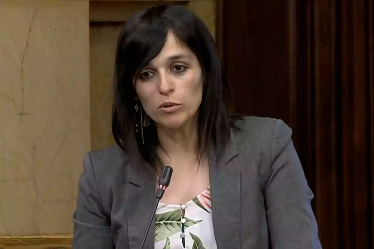 Una dona amb cabell fosc i jaqueta grisa parla davant d'un micròfon en un entorn formal.