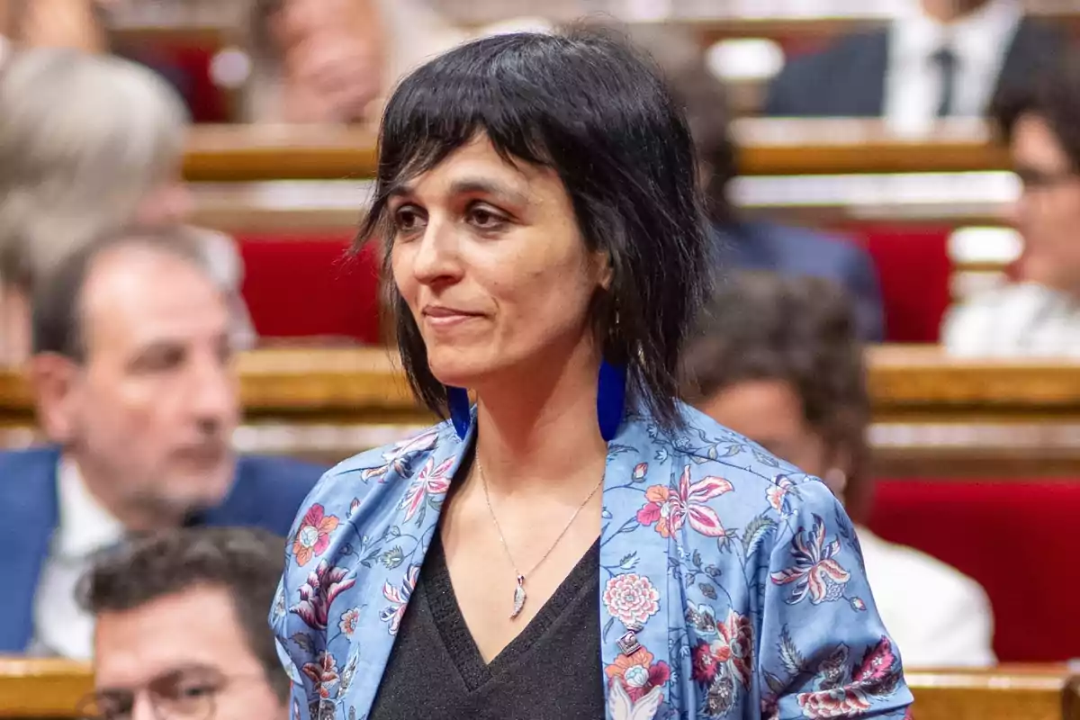 Una dona amb cabell curt i fosc, usant una jaqueta blava amb estampat floral i arracades blaves, està dreta en el que sembla ser una sala de reunions o un parlament, amb diverses persones desenfocades al fons.