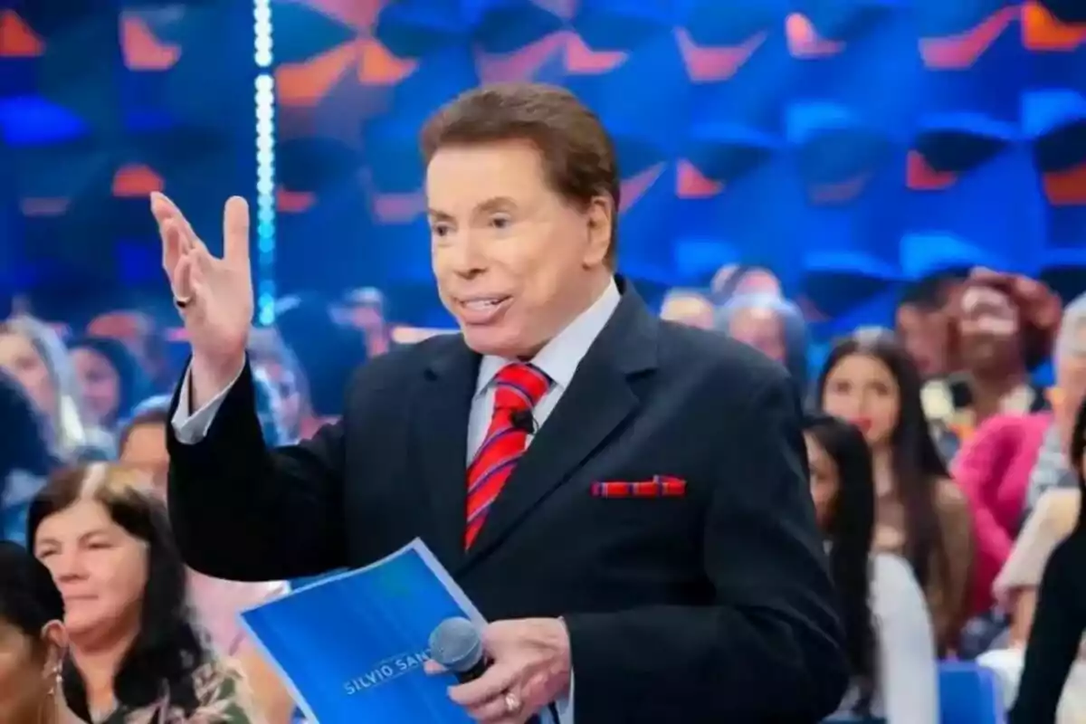 Fotografia de Silvio Santos en vestit i corbata vermella sostenint un micròfon i un document en un escenari amb una audiència al fons.