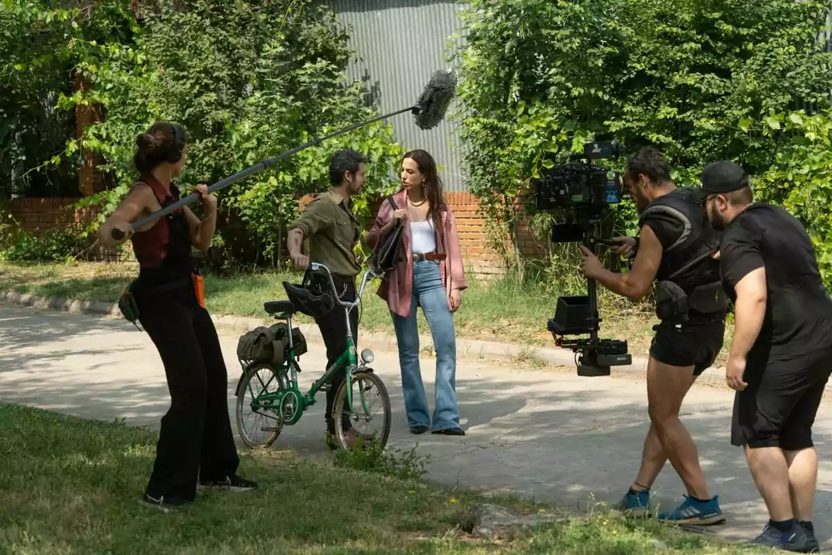 Equip de rodatge gravant una escena a l’aire lliure on una dona i un home amb bicicleta conversen mentre una persona sosté una perxa de micròfon i dos càmeres els enregistren a ‘Sense gluten’. Equip de rodatge gravant una escena a l’aire lliure on una dona i un home amb bicicleta conversen mentre una persona sosté una perxa de micròfon i dos càmeres els enregistren a ‘Sense gluten’.