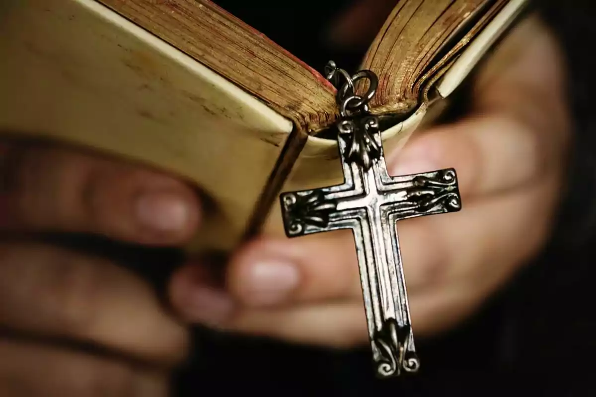 Manos sosteniendo un libro antiguo con un crucifijo de metal colgando.