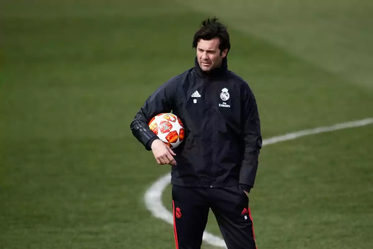A man in a black jacket and sweatpants holds a soccer ball on a training field. A man in a black jacket and sweatpants holds a soccer ball on a training field.