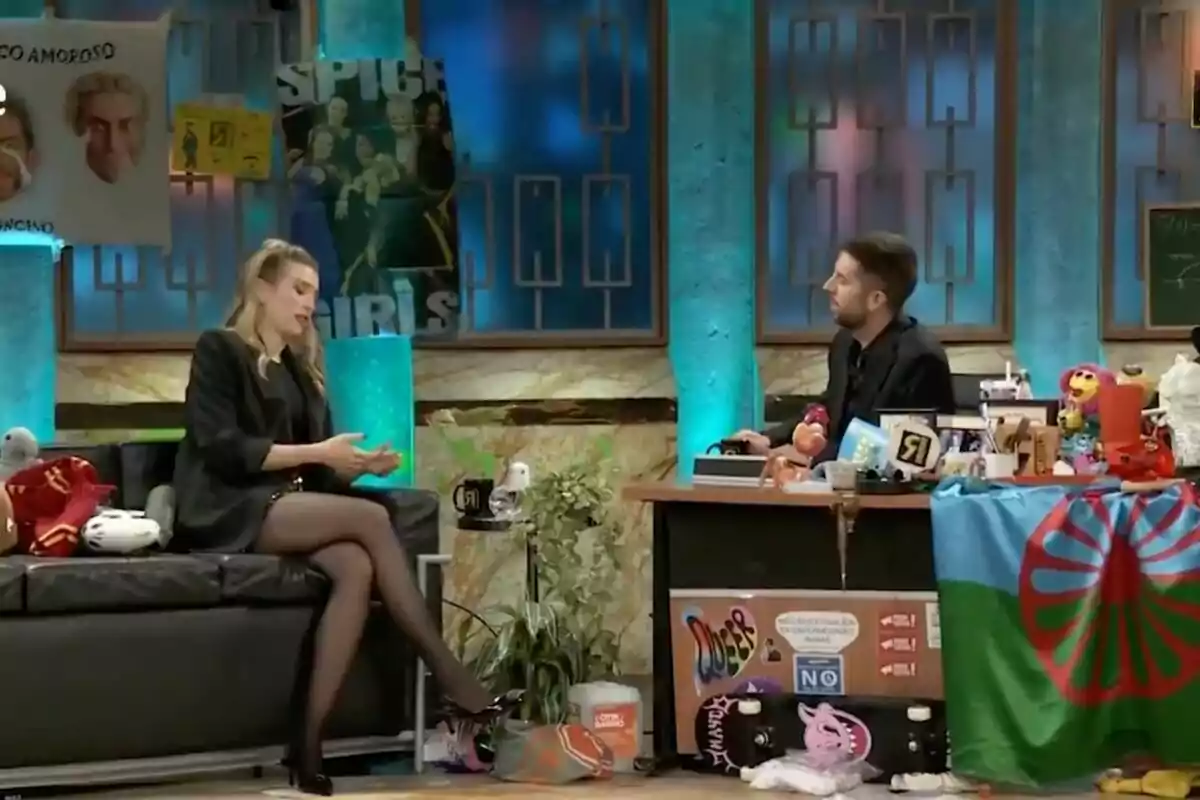 David Broncano y Eva Soriano conversan en un set de televisión decorado con carteles y objetos variados, mientras ella está sentada en un sofá y él detrás de un escritorio lleno de artículos diversos en 'La Revuelta'.