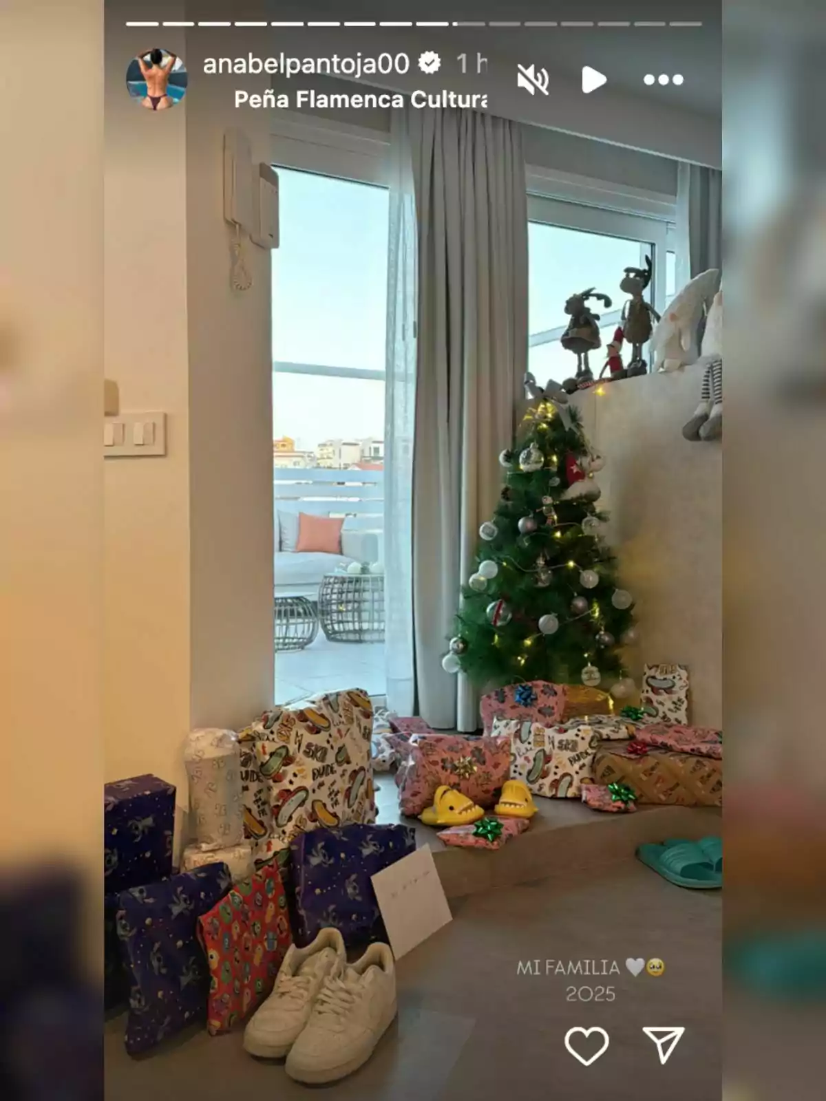 Una sala decorada para Navidad con un árbol pequeño adornado y varios regalos envueltos a su alrededor, junto a una ventana que deja ver el exterior.