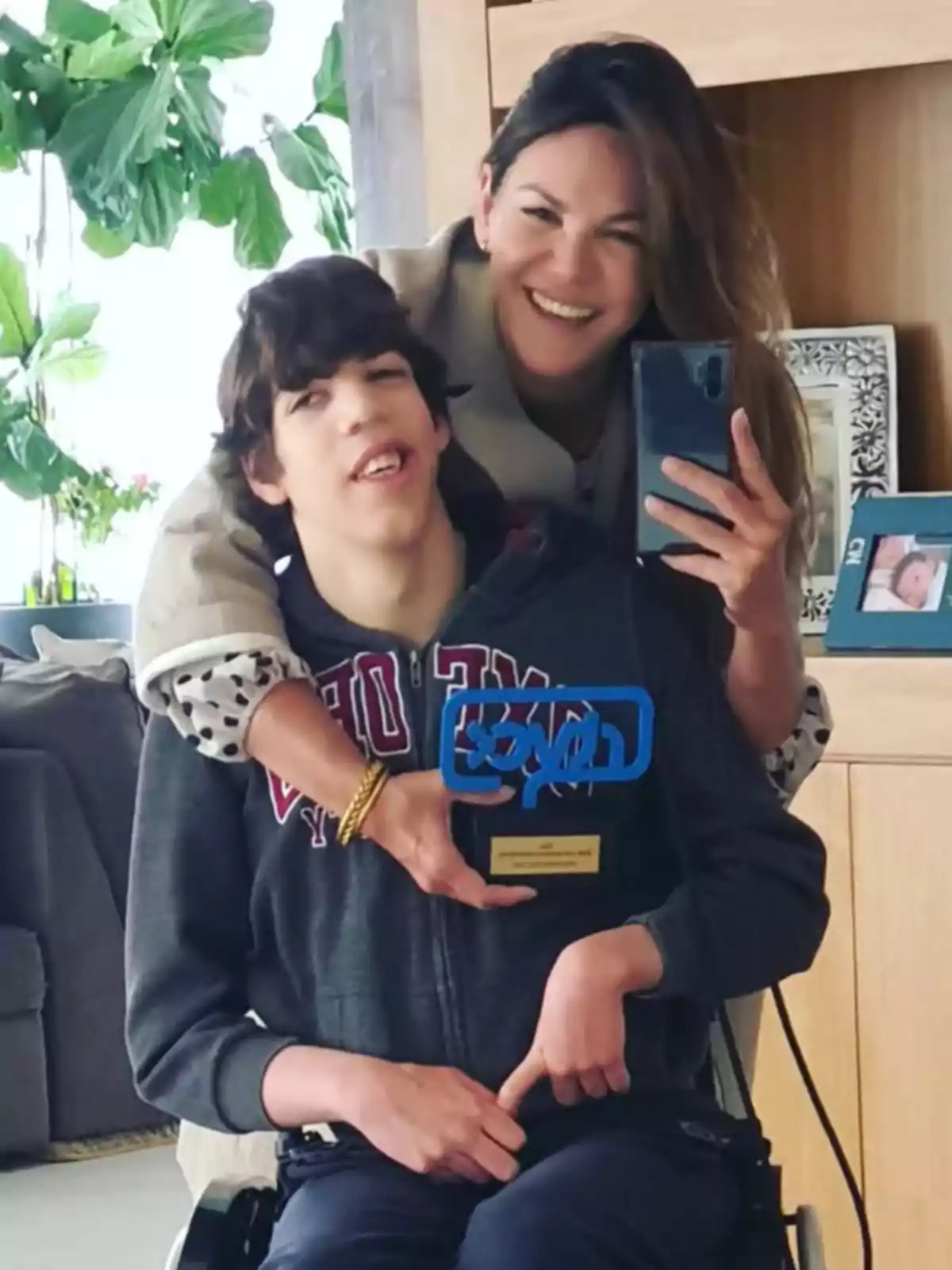 Fabiola Martínez, smiling, hugs Kike Osborne in a wheelchair as they take a selfie in front of a mirror; both seem happy and are in a room with plants and photos in the background.