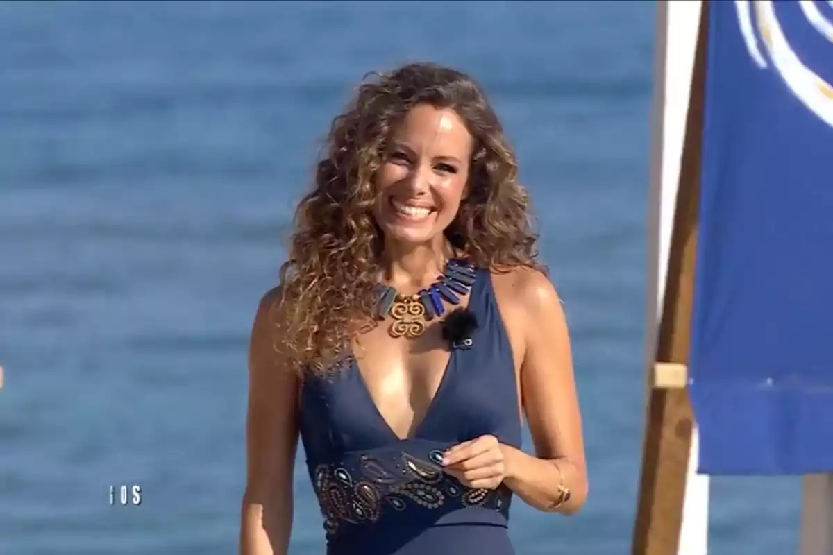 Una mujer sonriente con cabello rizado y un vestido azul oscuro con un collar llamativo, de pie frente al mar.