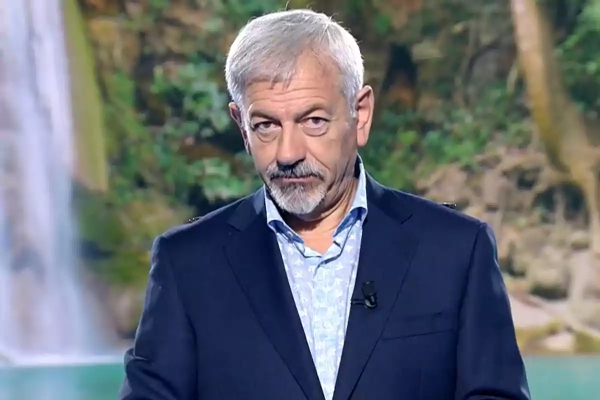 Carlos Sobera, vestit amb vestit fosc i camisa clara, està dret davant d'un fons natural amb vegetació i una cascada. Captura de 'Supervivientes'. Carlos Sobera, vestit amb vestit fosc i camisa clara, està dret davant d'un fons natural amb vegetació i una cascada. Captura de 'Supervivientes'.