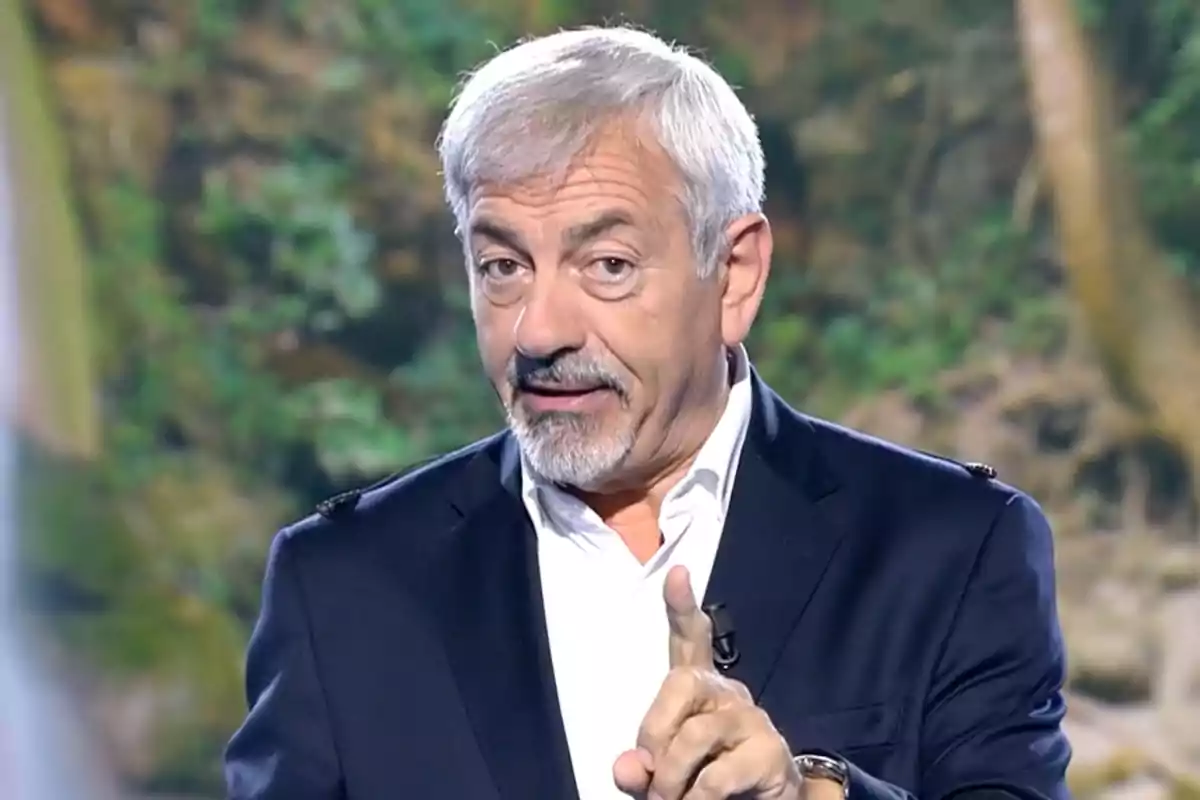 Carlos Sobera està parlant i gesticulant amb el dit índex aixecat, vestint un vestit fosc i camisa blanca, amb un fons de natura desenfocat. Captura de 'Supervivientes'.