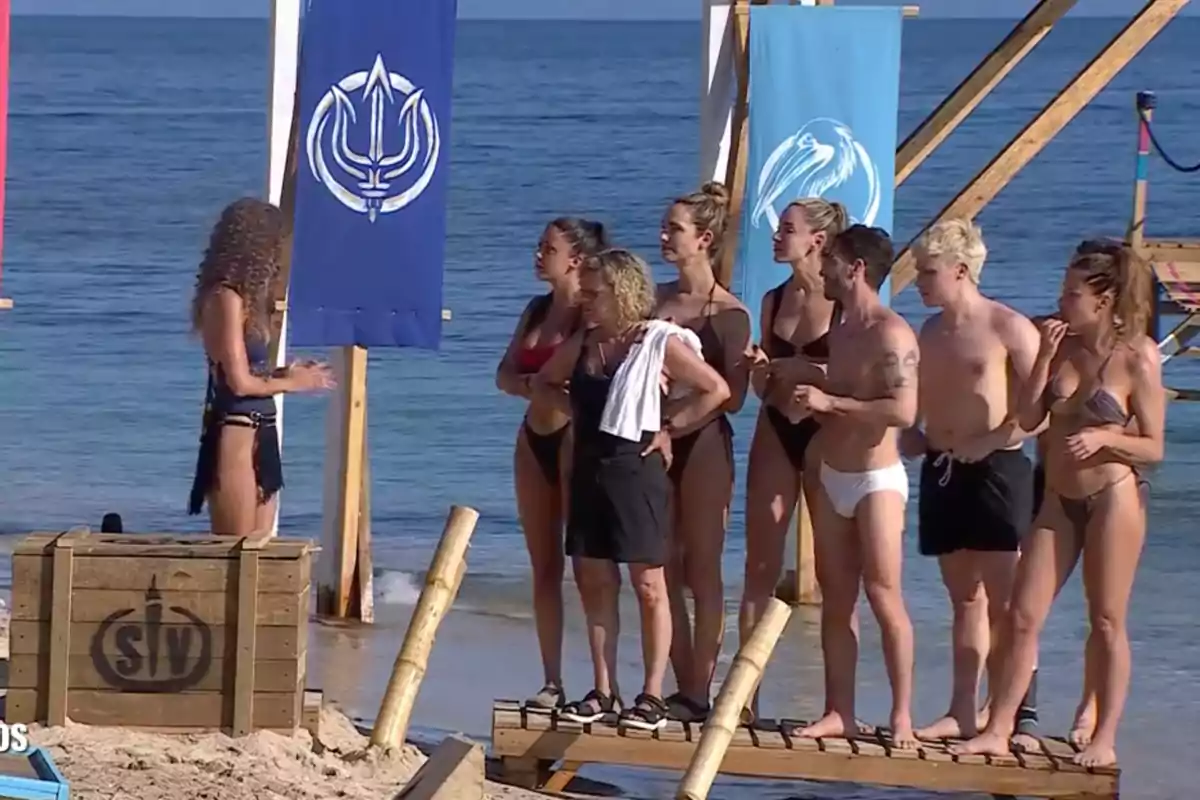 A group of people in swimsuits is standing on the beach in front of a woman who seems to be speaking, with blue flags in the background and the sea behind. A group of people in swimsuits is standing on the beach in front of a woman who seems to be speaking, with blue flags in the background and the sea behind.