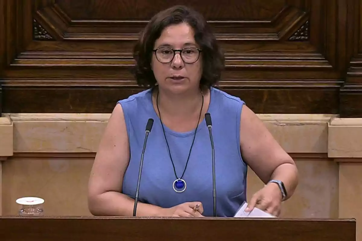 Short-haired woman with glasses speaking at a podium with two microphones in an institutional setting Short-haired woman with glasses speaking at a podium with two microphones in an institutional setting