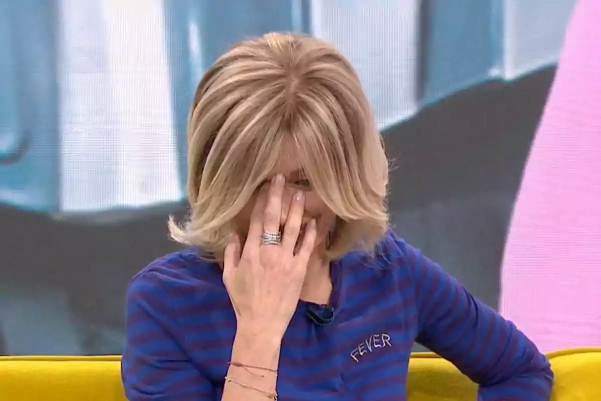 Susanna Grisoo amb cabell ros es cobreix la cara amb la mà mentre està asseguda en un sofà groc i porta una samarreta blava amb ratlles a 'Espejo Público'.