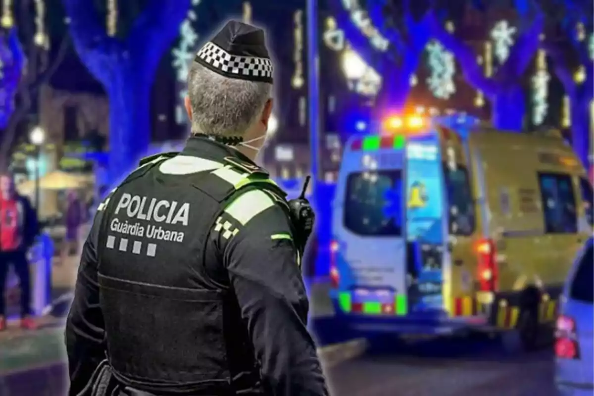 Un agente de la Guardia Urbana de espaldas observa una ambulancia con luces encendidas en una calle iluminada por decoraciones nocturnas.