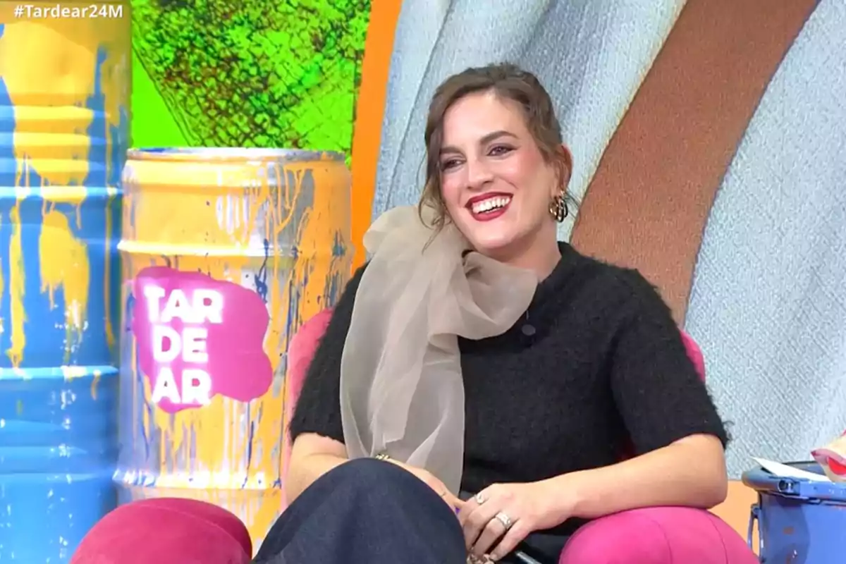 Leticia Requejo, smiling, is sitting on a pink armchair in a colorful television set with barrels painted yellow and blue. Capture from 'TardeAR'.