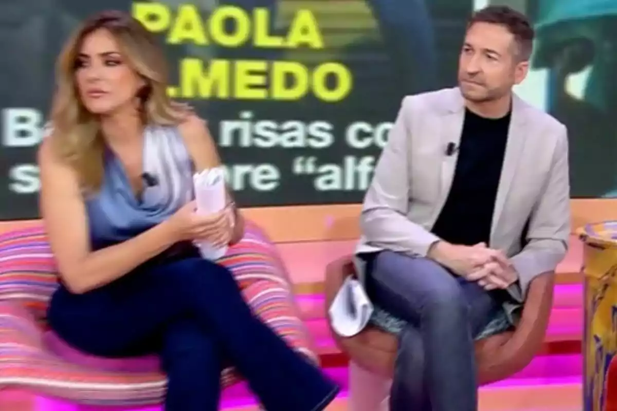 Frank Blanco i Verónica Dulanto asseguts a 'Tardear' acolorit amb un fons que mostra lletres grogues i blanques Frank Blanco i Verónica Dulanto asseguts a 'Tardear' acolorit amb un fons que mostra lletres grogues i blanques