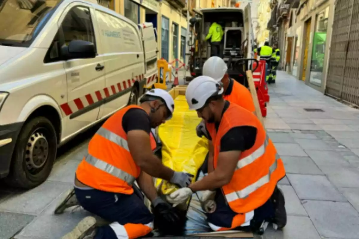 Treballadors de la construcció amb armilles taronges i cascos blancs fan tasques en un carrer al costat d'una furgoneta blanca.