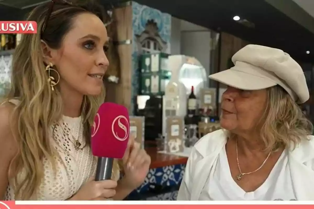 Una reportera sosté un micròfon rosa de Socialité mentre entrevista Terelu Campos, de cabell ros i gorra beix, en un lloc tancat. Una reportera sosté un micròfon rosa de Socialité mentre entrevista Terelu Campos, de cabell ros i gorra beix, en un lloc tancat.