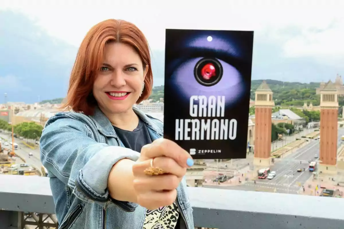 Fotografia de Teresa Colomina somrient amb cabells vermells sosté un cartell del programa "Gran Hermano" amb Barcelona de fons.