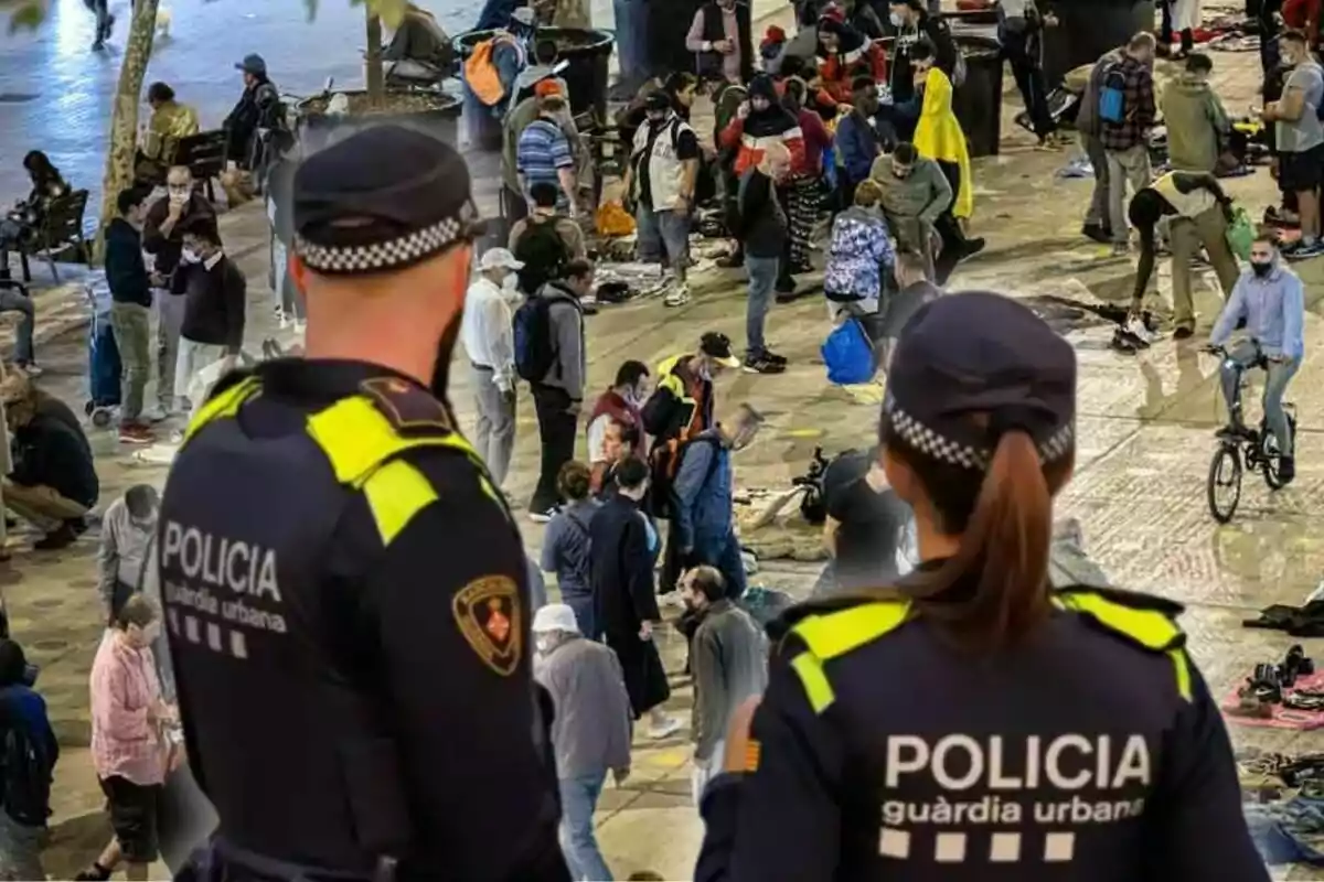 Dos agents de la policia observen una concorreguda plaça on hi ha moltes persones reunides, algunes caminant i d'altres amb bicicleta.