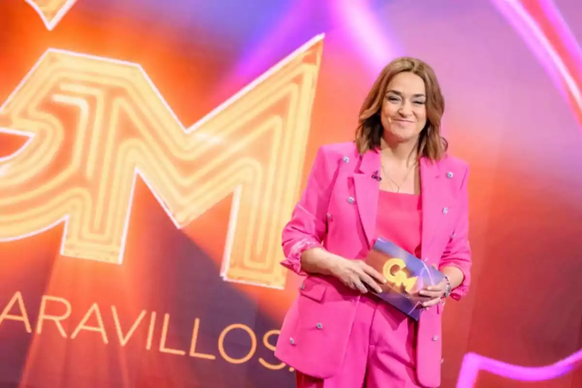 Toñi Moreno amb vestit rosa sostenint una targeta en un escenari amb fons acolorit a Gent Meravellosa.
