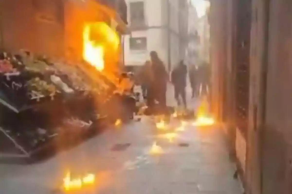 Una escena d'un carrer estret amb flames i fum sortint d'un edifici mentre diverses persones caminen a prop.