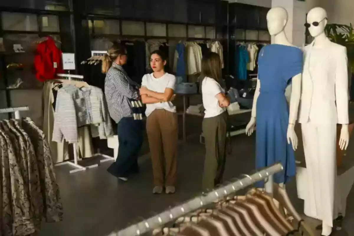 Persones observen roba en una botiga de moda, amb maniquins vestits al davant i penjadors plens de peces al fons. Persones observen roba en una botiga de moda, amb maniquins vestits al davant i penjadors plens de peces al fons.