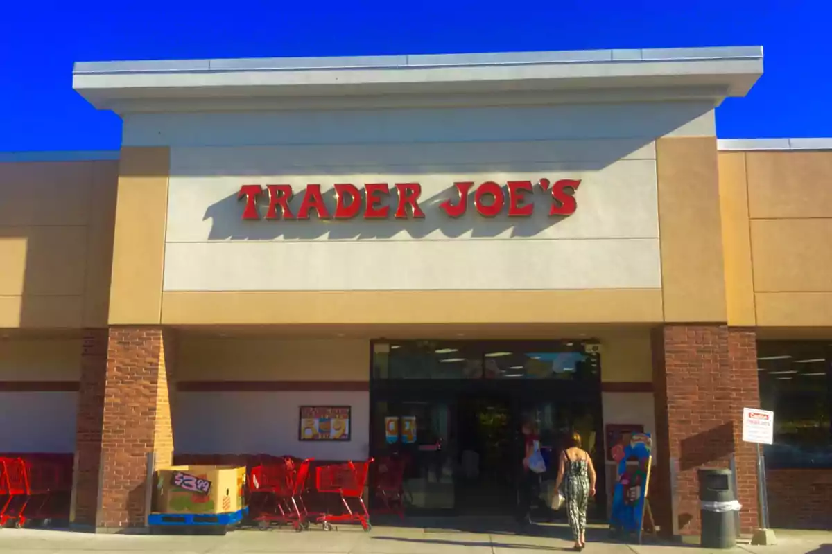 Fachada de una tienda con el letrero "Trader Joe's" en letras rojas, con carros de supermercado rojos y personas entrando al establecimiento.