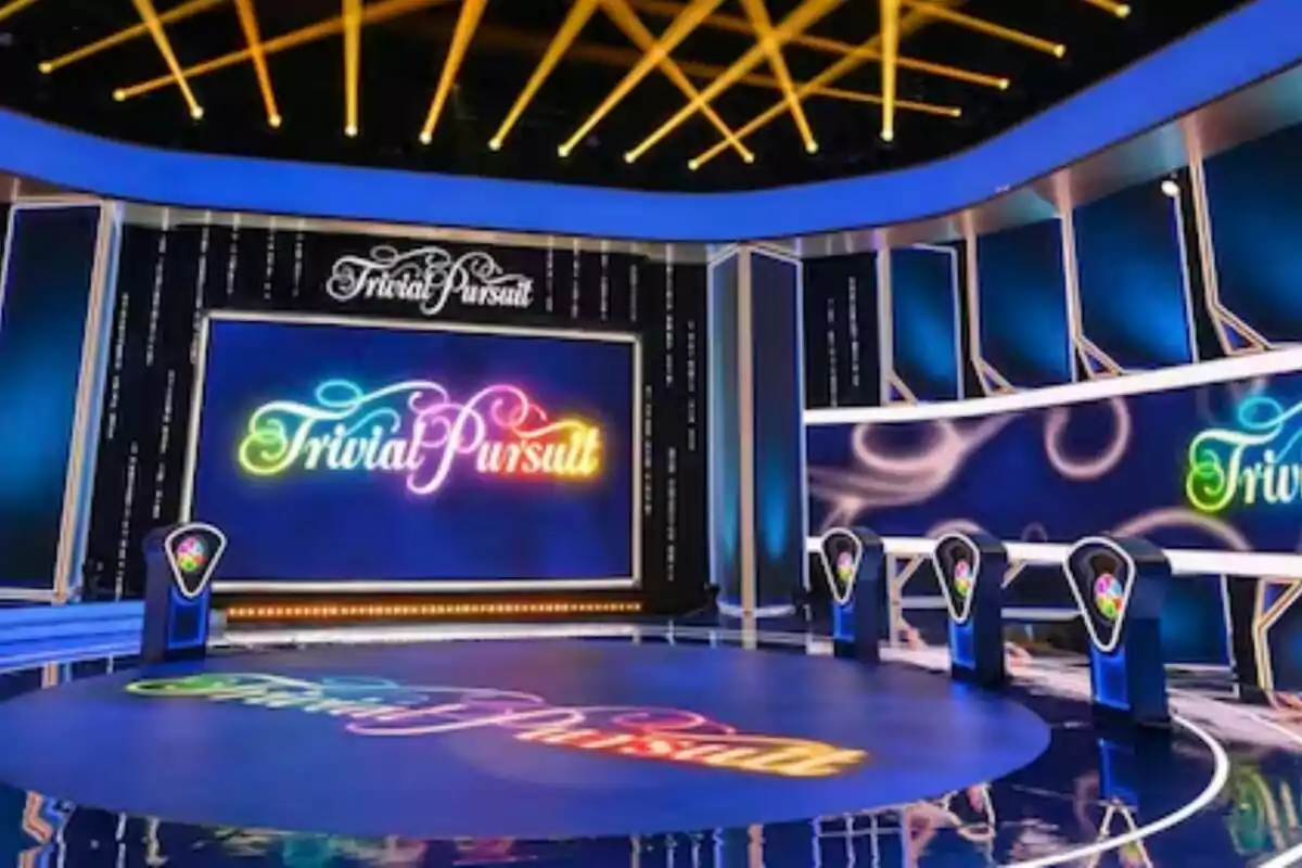 Escenari acolorit d’un programa de concursos amb el logotip de Trivial Pursuit a pantalles grans i faristols il·luminats per als participants