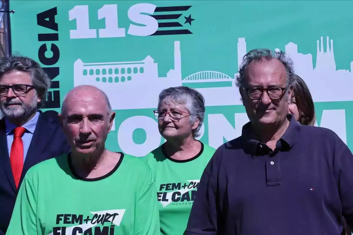 Un grup de persones posant davant d'un cartell verd que diu “11S” i “Fem + Curt El Camí”.