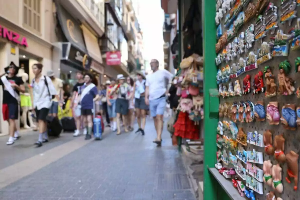 Un grup de persones camina per un carrer comercial ple de botigues, mentre que en primer pla es veuen imants de record en un lloc.