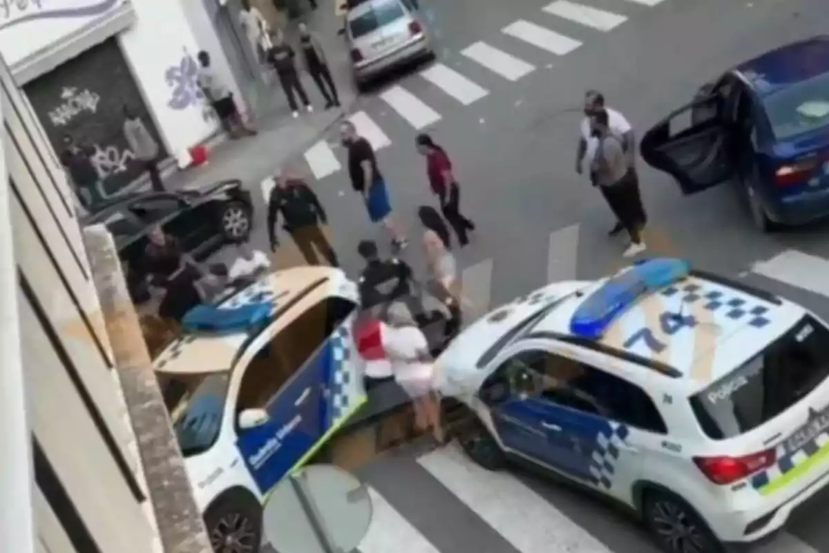 A la imatge s'observa una escena urbana amb dos vehicles policials i diverses persones al voltant, alguns sembla que estan interactuant amb els agents de policia.