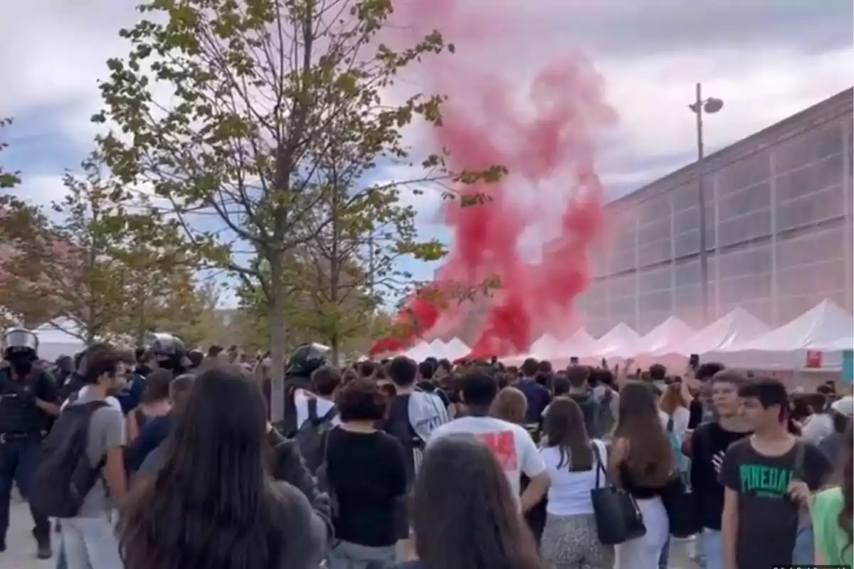 Una multitud de persones es reuneixen a l'aire lliure, amb fum vermell elevant-se al fons i diverses carpes blanques alineades al llarg d'un edifici.