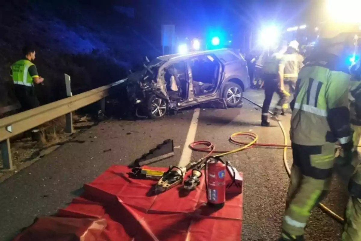 Accident de trànsit nocturn amb un cotxe greument danyat al costat de la tanca de seguretat, bombers i equip de rescat treballant a l’escena amb eines i extintor en primer pla