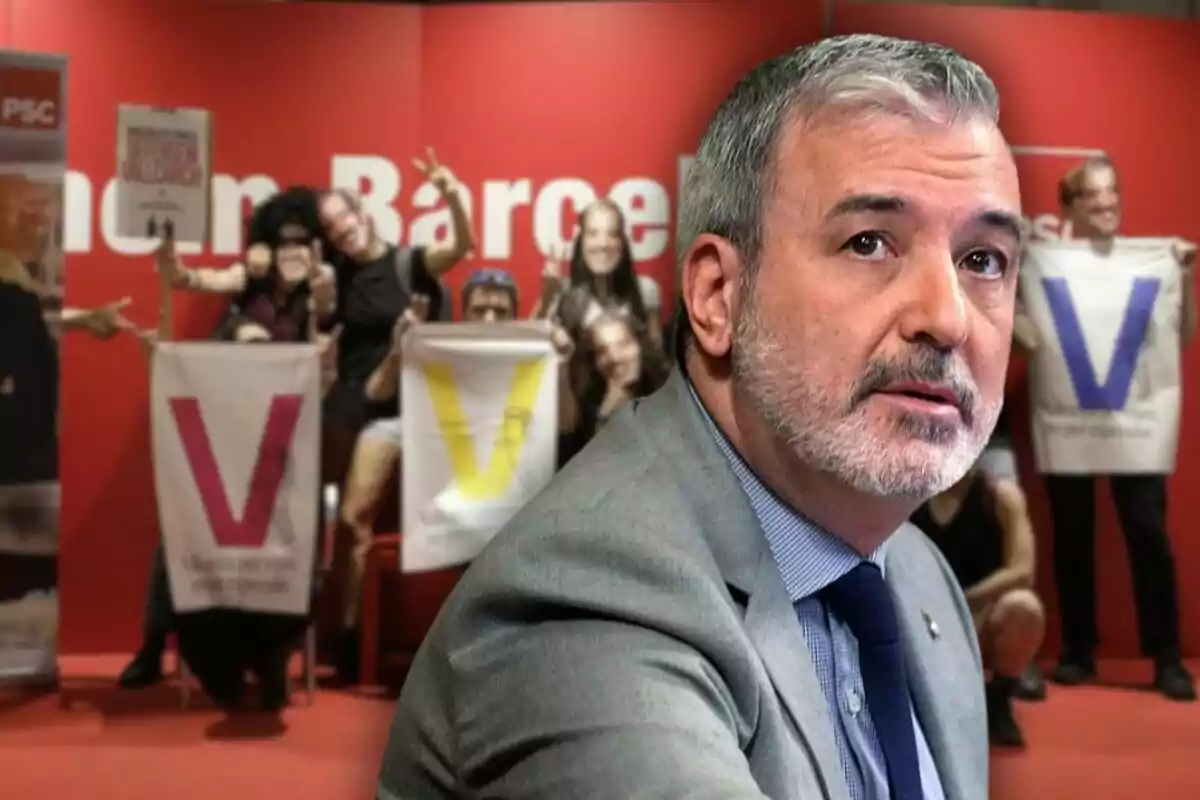 Home de cabells canosos i barba amb vestit i corbata en primer pla, al fons persones sostenen pancartes amb lletres grans i colorides sobre un fons vermell.