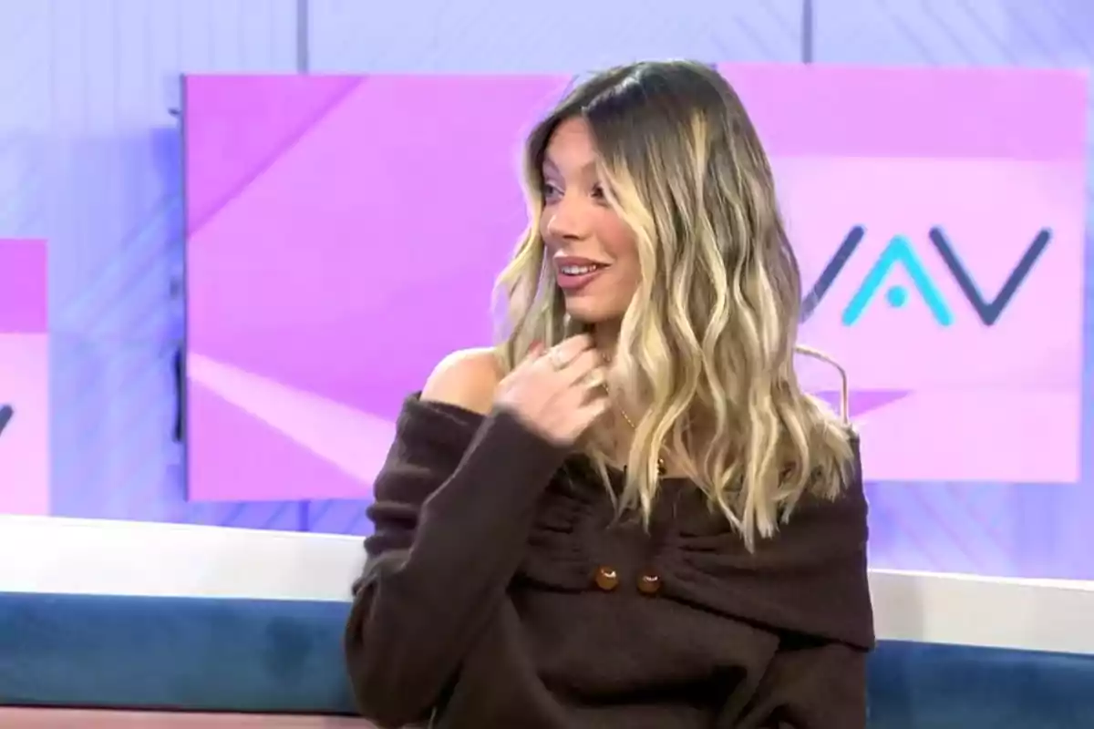 Alejandra Rubio with wavy blonde hair and a dark brown sweater is sitting on a television set with pink and purple screens in the background. Capture from 'Vamos a Ver'