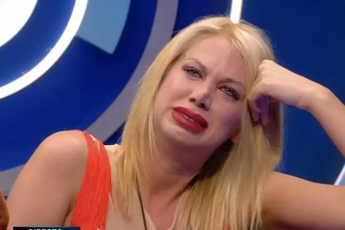 Vanessa Bouza rubia con expresión de tristeza y lágrimas en los ojos, apoyando su cabeza en su mano, con un fondo azul en GH DÚO.