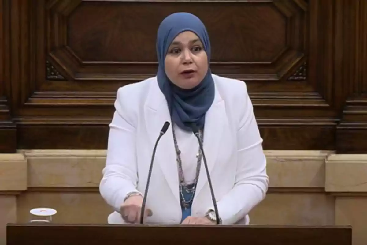 Una dona amb hijab blau i vestit blanc parla en un podi amb micròfons davant d'un fons de fusta.