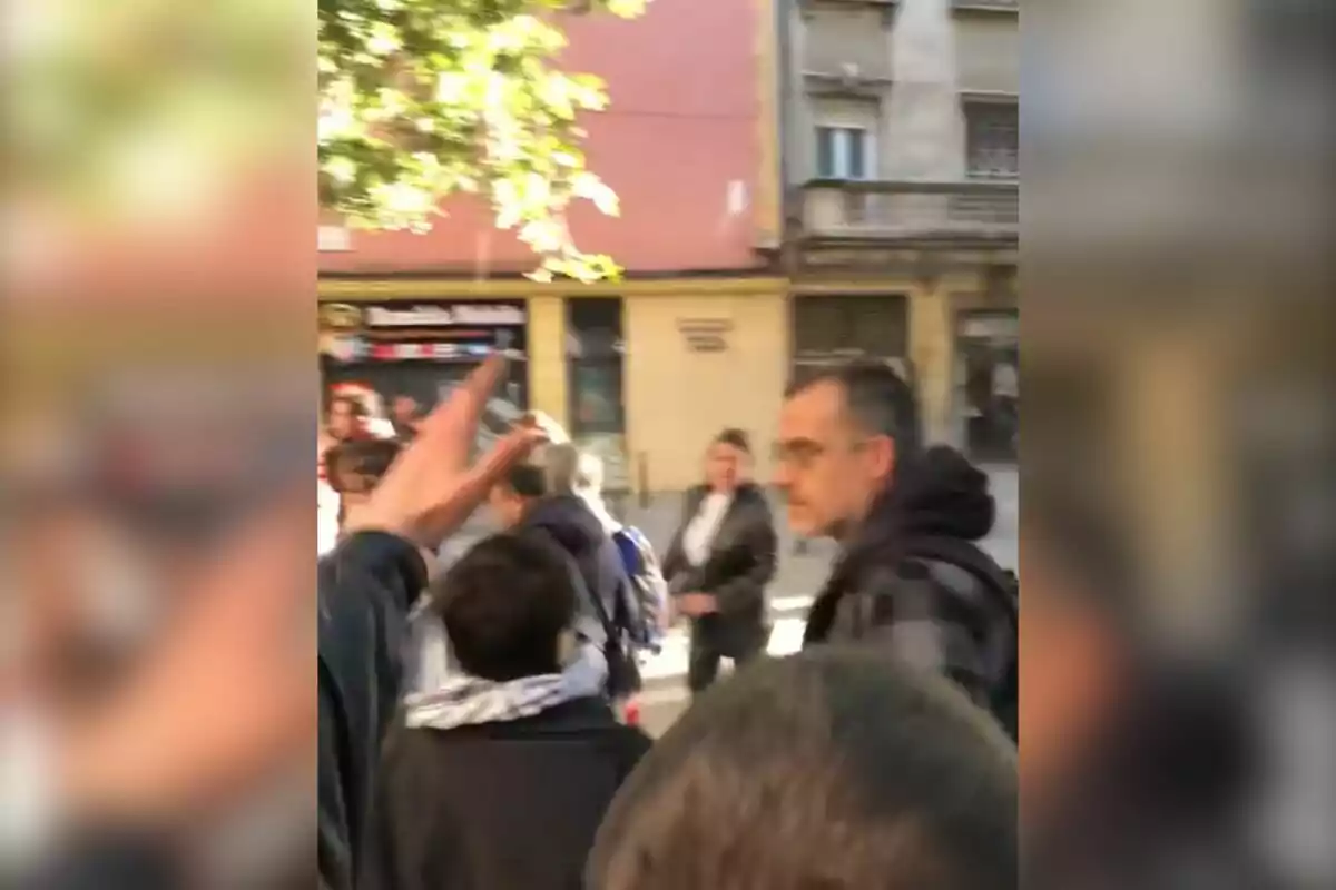 Un grup de persones caminen per un carrer amb edificis al fons i un arbre que projecta ombra.