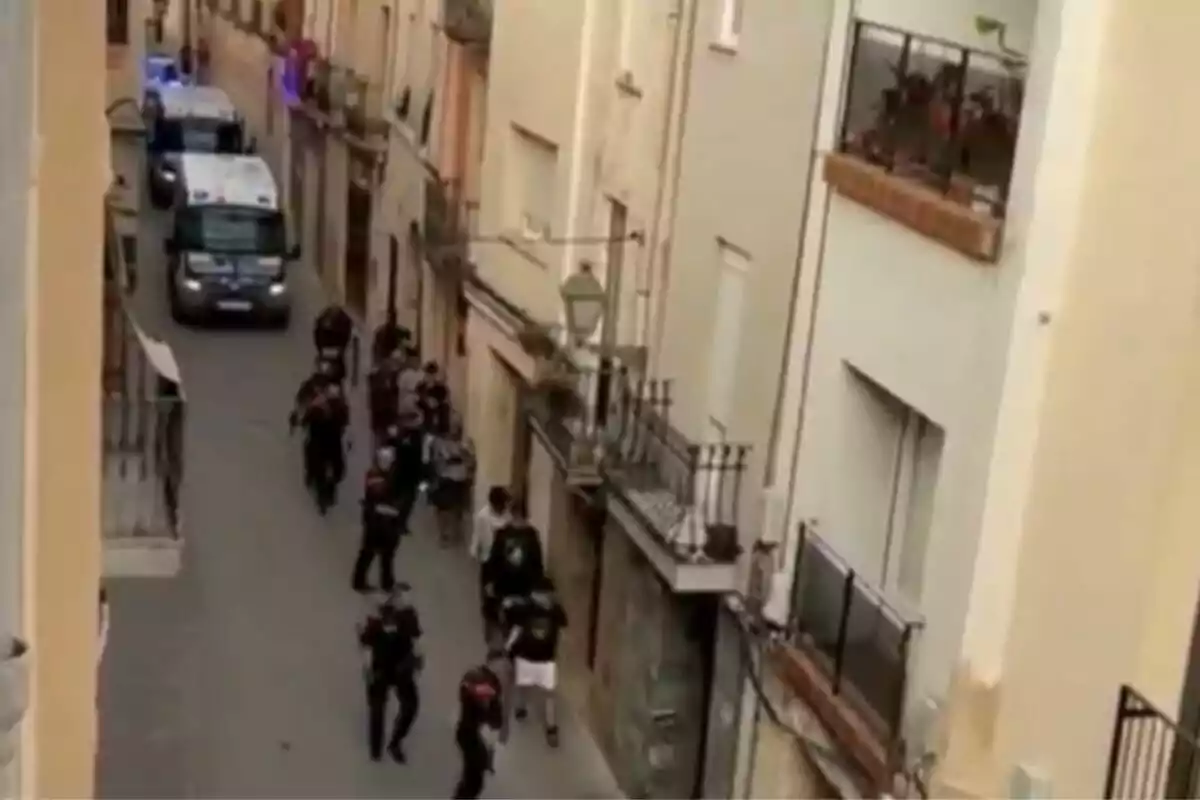 Un grup de persones camina per un carrer estret flanquejat per edificis alts, mentre diversos vehicles policials estan estacionats al fons.