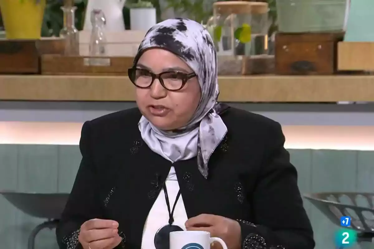 Una persona amb ulleres i un mocador al cap està parlant en un entorn que sembla ser un estudi de televisió o una cuina, amb diversos objectes i plantes al fons.