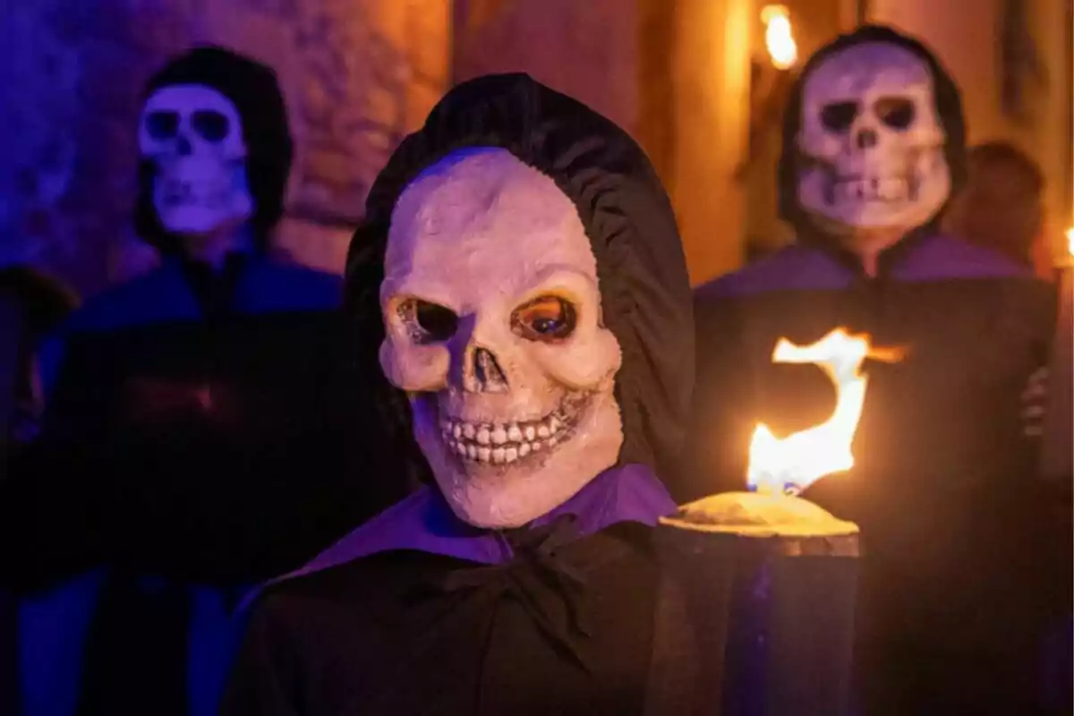 Persones amb màscares de calavera i túniques fosques sostenen torxes enceses en un ambient il·luminat per llums càlides. Persones amb màscares de calavera i túniques fosques sostenen torxes enceses en un ambient il·luminat per llums càlides.