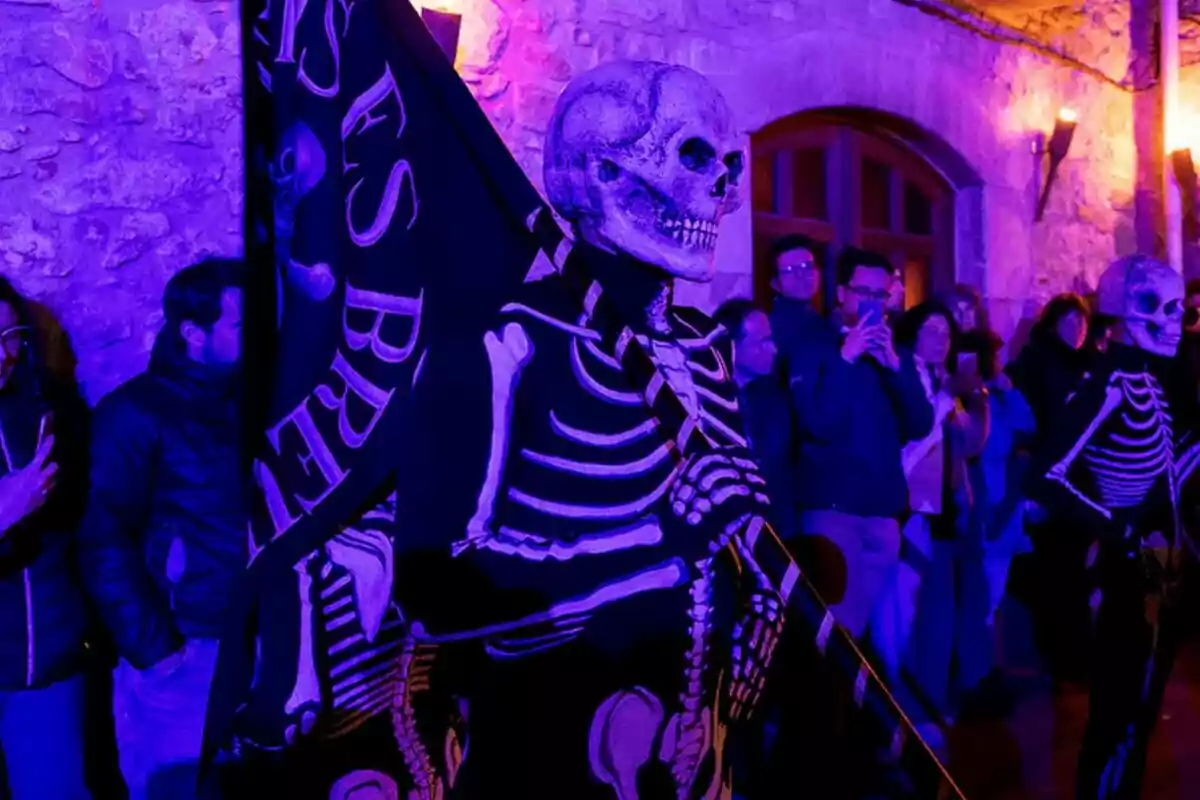 Persones disfressades d'esquelets sostenen una bandera en un esdeveniment nocturn amb il·luminació porpra, mentre el públic observa. Persones disfressades d'esquelets sostenen una bandera en un esdeveniment nocturn amb il·luminació porpra, mentre el públic observa.