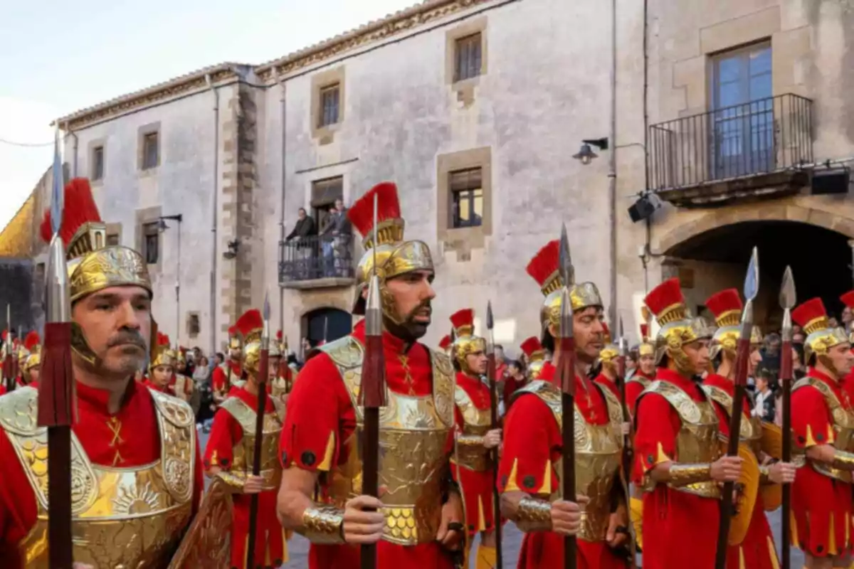 Grup de persones vestides com a soldats romans amb armadures daurades i túniques vermelles en un carrer davant d'un edifici antic. Grup de persones vestides com a soldats romans amb armadures daurades i túniques vermelles en un carrer davant d'un edifici antic.