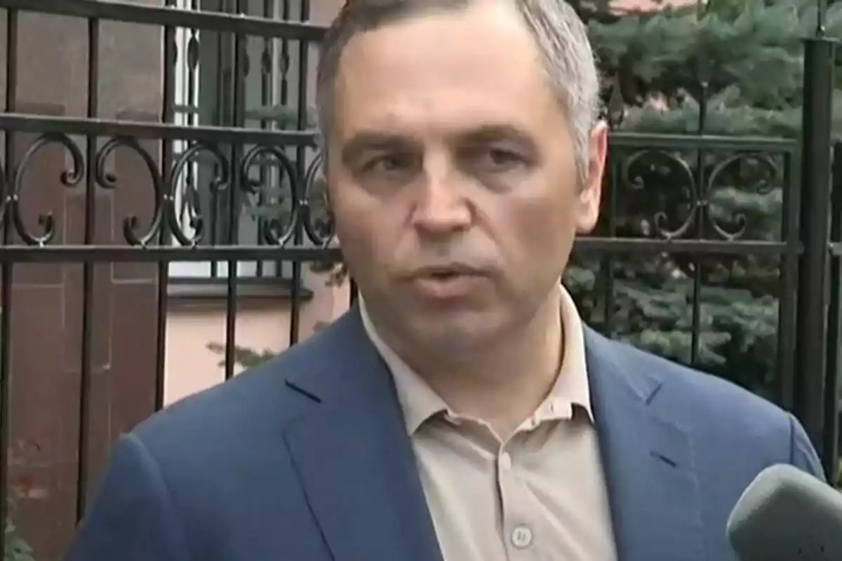 Un home amb jaqueta blava i camisa beix parla davant d'una reixa metàl·lica mentre sosté un micròfon. Un home amb jaqueta blava i camisa beix parla davant d'una reixa metàl·lica mentre sosté un micròfon.
