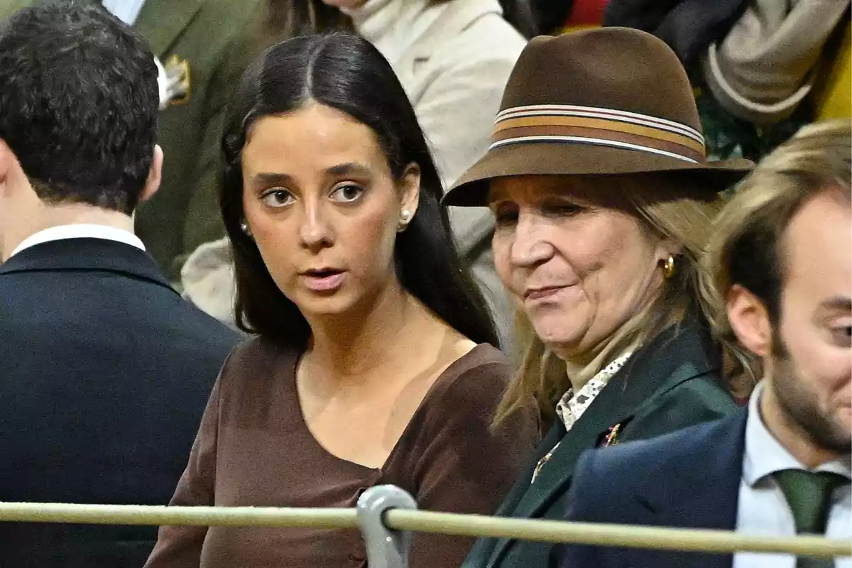 Victoria Federica y la infanta Elena sentadas en un evento, una mujer joven con cabello oscuro y otra mujer con sombrero marrón.