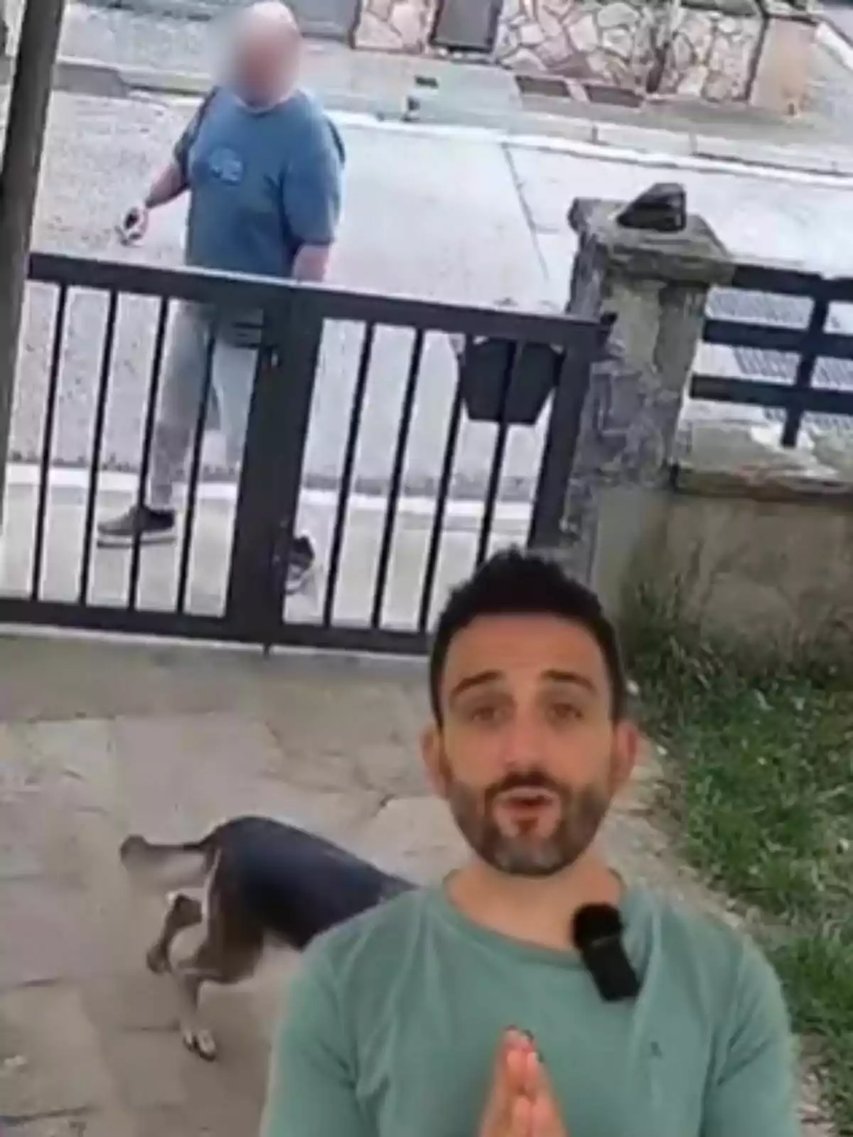 Un home amb samarreta blava camina per la vorera mentre un gos és al pati darrere d'una reixa i un home amb samarreta verda apareix en primer pla. Un home amb samarreta blava camina per la vorera mentre un gos és al pati darrere d'una reixa i un home amb samarreta verda apareix en primer pla.