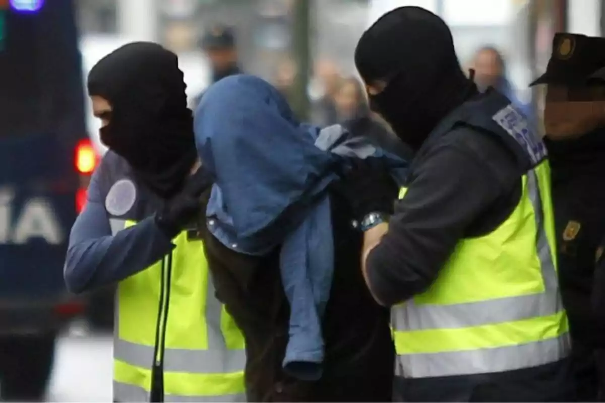 People with reflective vests and balaclavas escort a hooded individual in an urban setting.